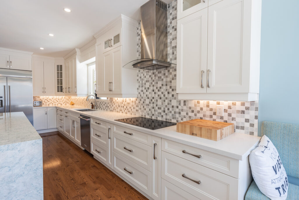 Bright transitional kitchen with custom white shaker cabinets, oversized quartz island, glass display cabinets, mosaic backsplash, and stainless steel appliances.