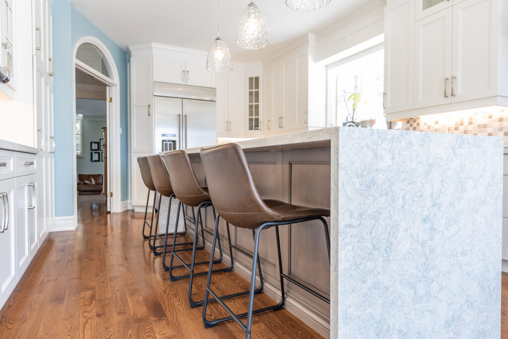 Bright transitional kitchen with custom white shaker cabinets, oversized quartz island, glass display cabinets, mosaic backsplash, and stainless steel appliances.
