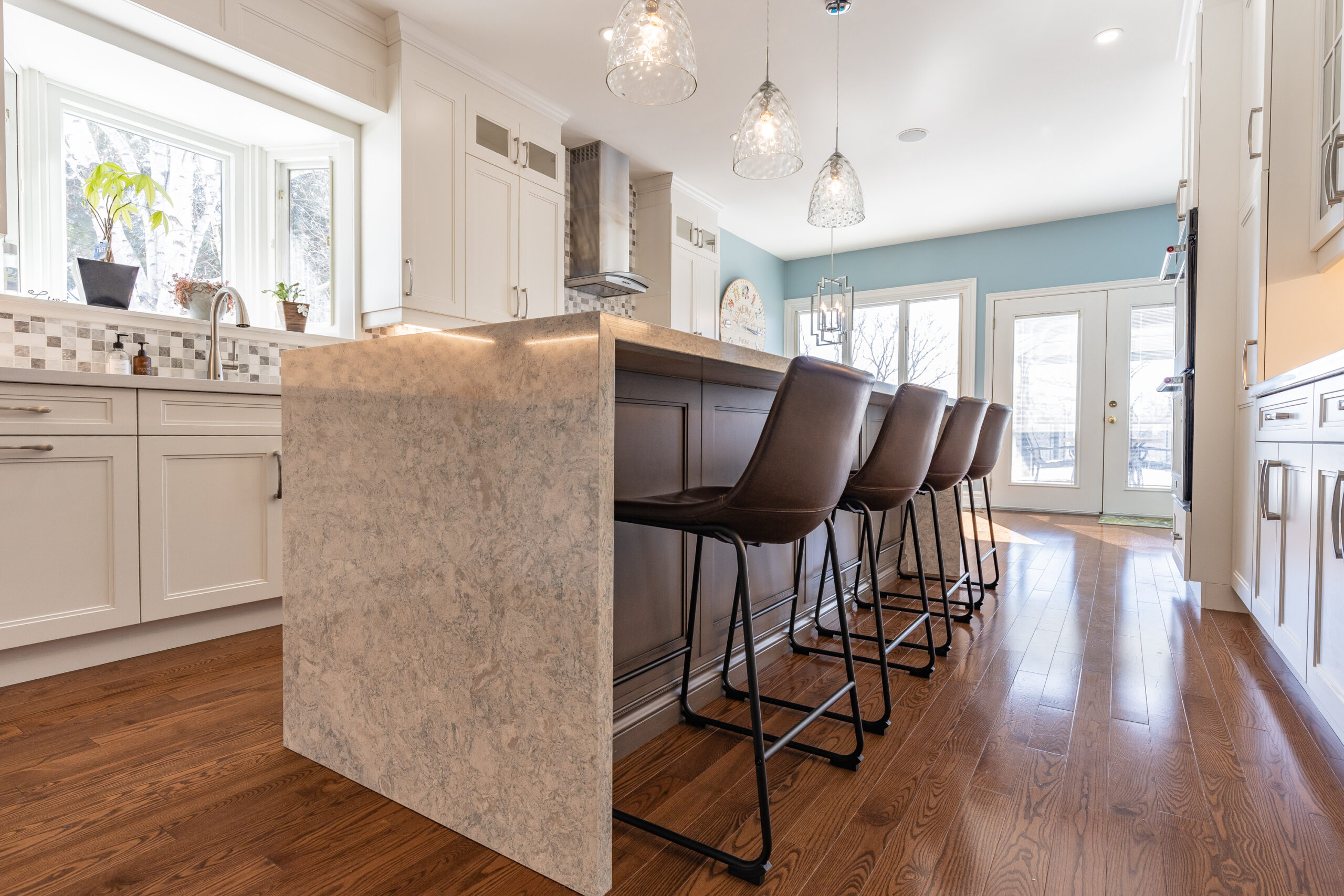 Bright transitional kitchen with custom white shaker cabinets, oversized quartz waterfall island, glass display cabinets, mosaic backsplash, and stainless steel appliances.