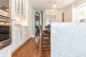 Bright transitional kitchen with custom white shaker cabinets, oversized quartz island, glass display cabinets, mosaic backsplash, and stainless steel appliances.