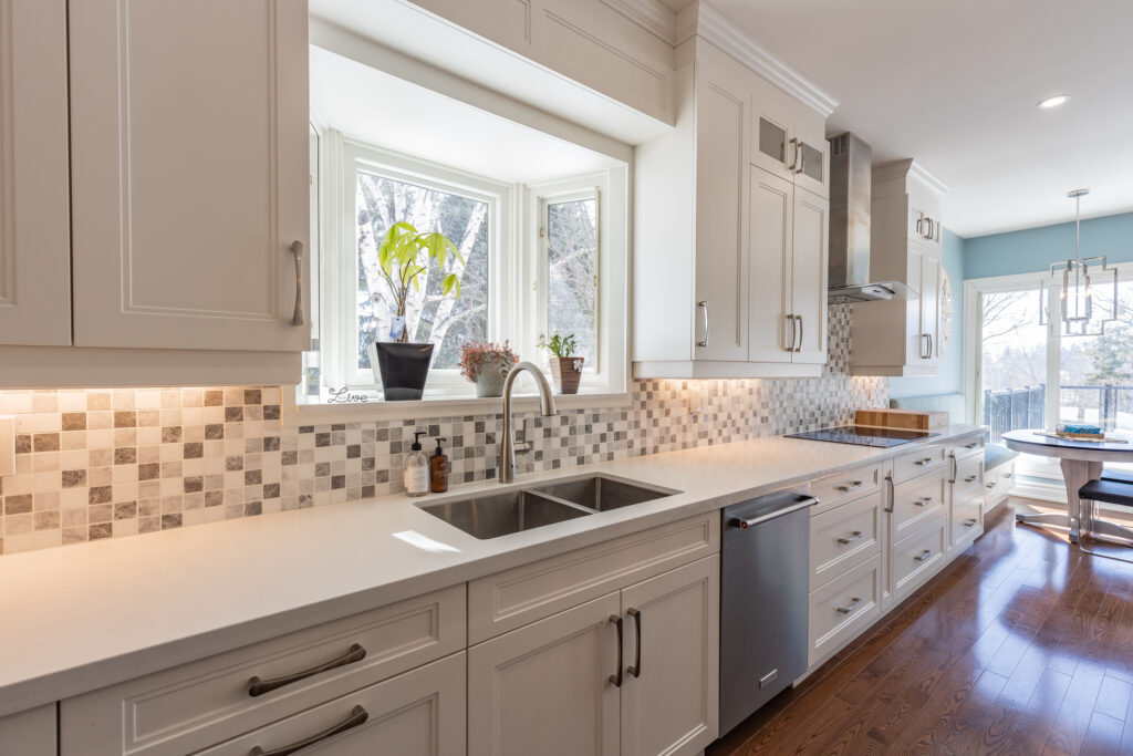 Bright transitional kitchen with custom white shaker cabinets, oversized quartz island, glass display cabinets, mosaic backsplash, and stainless steel appliances.