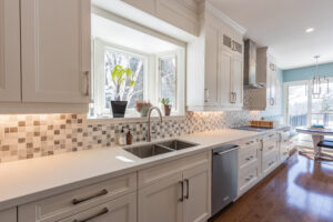 Bright transitional kitchen with custom white shaker cabinets, oversized quartz island, glass display cabinets, mosaic backsplash, and stainless steel appliances.
