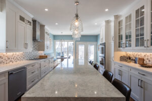 Bright transitional kitchen with custom white shaker cabinets, oversized quartz island, glass display cabinets, mosaic backsplash, and stainless steel appliances.
