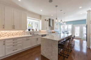 Bright transitional kitchen with custom white shaker cabinets, oversized quartz island, glass display cabinets, mosaic backsplash, and stainless steel appliances.