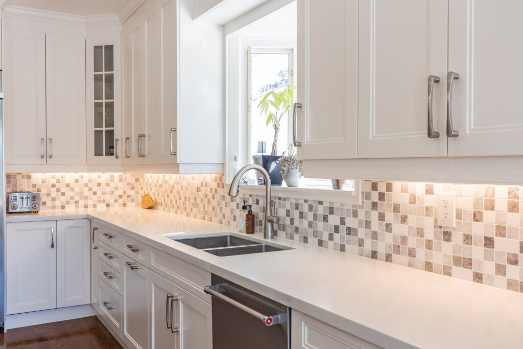 Bright transitional kitchen with custom white shaker cabinets, oversized quartz island, glass display cabinets, mosaic backsplash, and stainless steel appliances.