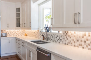Bright transitional kitchen with custom white shaker cabinets, oversized quartz island, glass display cabinets, mosaic backsplash, and stainless steel appliances.