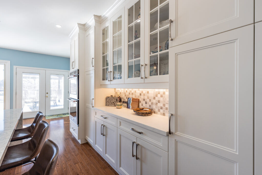 Bright transitional kitchen with custom white shaker cabinets, oversized quartz island, glass display cabinets, mosaic backsplash, and stainless steel appliances.