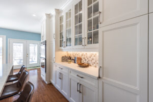 Bright transitional kitchen with custom white shaker cabinets, oversized quartz island, glass display cabinets, mosaic backsplash, and stainless steel appliances.