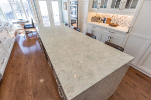Bright transitional kitchen with custom white shaker cabinets, oversized quartz island, glass display cabinets, mosaic backsplash, and stainless steel appliances.