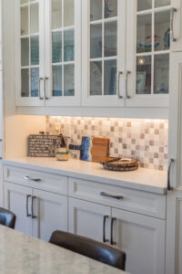 Bright transitional kitchen with custom white shaker cabinets, oversized quartz island, glass display cabinets, mosaic backsplash, and stainless steel appliances.