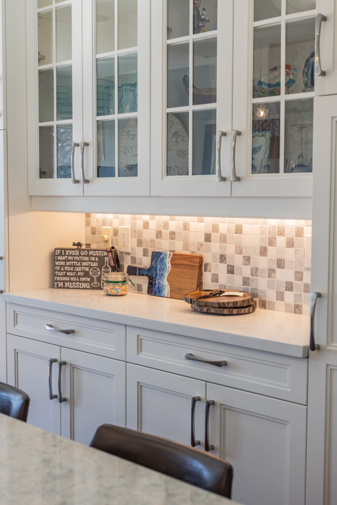 Bright transitional kitchen with custom white shaker cabinets, oversized quartz island, glass display cabinets, mosaic backsplash, and stainless steel appliances.