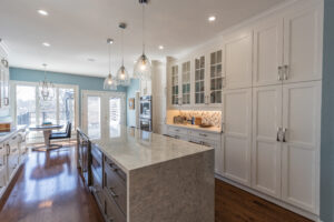 Bright transitional kitchen with custom white shaker cabinets, oversized quartz island, glass display cabinets, mosaic backsplash, and stainless steel appliances.