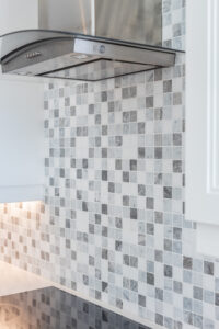 Bright transitional kitchen with custom white shaker cabinets, oversized quartz island, glass display cabinets, mosaic backsplash, and stainless steel appliances.