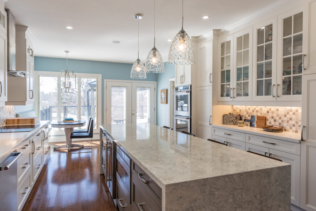 Bright transitional kitchen with custom white shaker cabinets, oversized quartz island, glass display cabinets, mosaic backsplash, and stainless steel appliances.