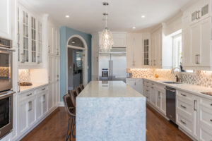 Bright transitional kitchen with custom white shaker cabinets, oversized quartz island, glass display cabinets, mosaic backsplash, and stainless steel appliances.