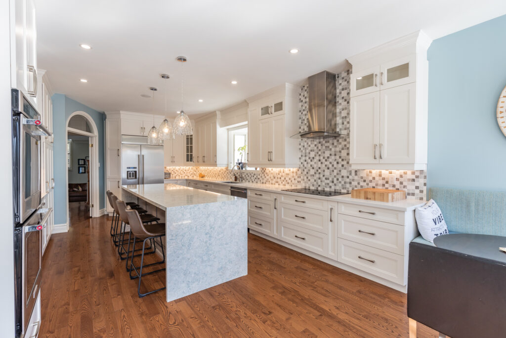 Bright transitional kitchen with custom white shaker cabinets, oversized quartz island, glass display cabinets, mosaic backsplash, and stainless steel appliances.