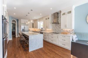 Bright transitional kitchen with custom white shaker cabinets, oversized quartz island, glass display cabinets, mosaic backsplash, and stainless steel appliances.