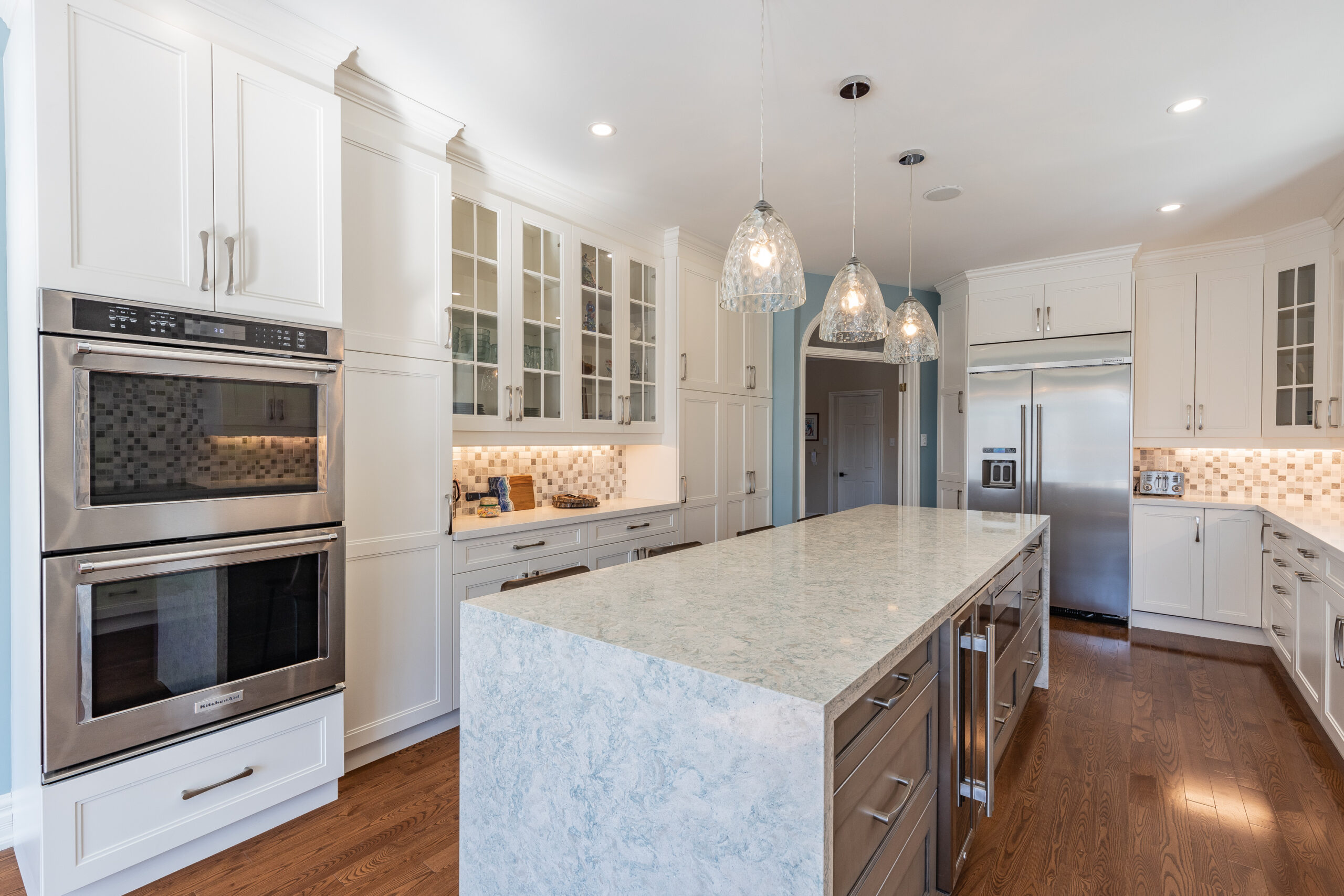 Bright transitional kitchen with custom white shaker cabinets, oversized quartz island, glass display cabinets, mosaic backsplash, and stainless steel appliances.