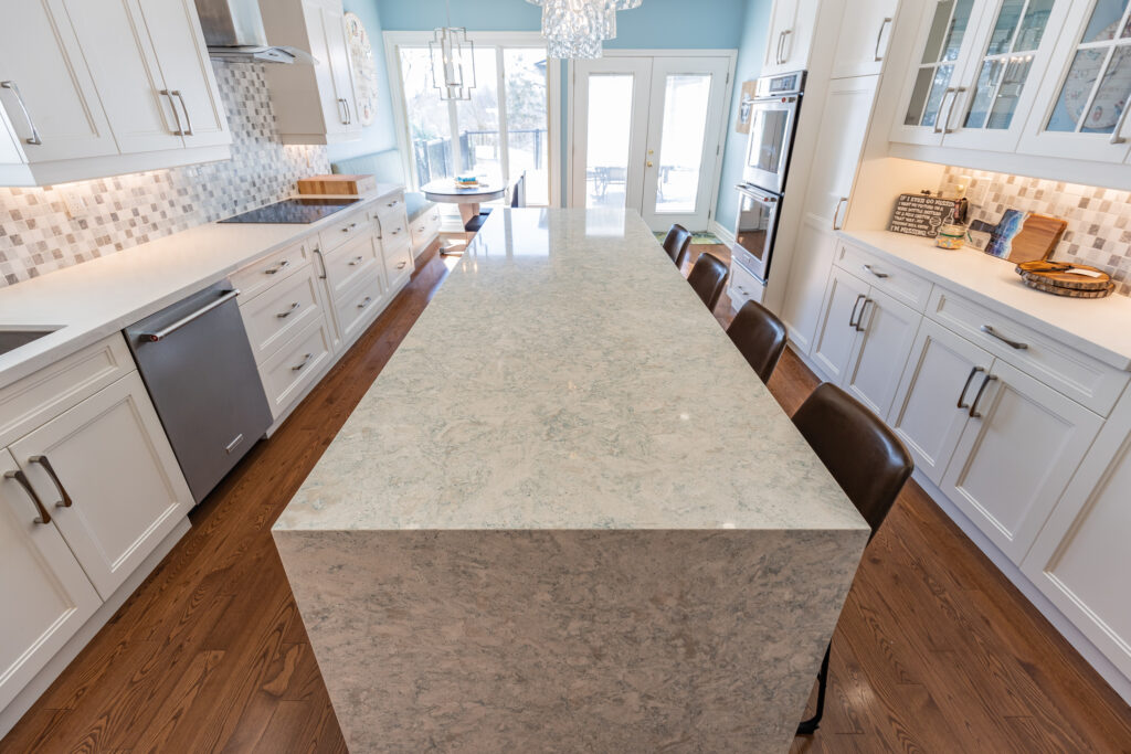 Bright transitional kitchen with custom white shaker cabinets, oversized quartz island, glass display cabinets, mosaic backsplash, and stainless steel appliances.