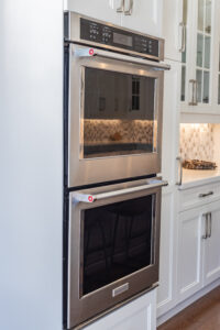 Bright transitional kitchen with custom white shaker cabinets, oversized quartz island, glass display cabinets, mosaic backsplash, and stainless steel appliances.